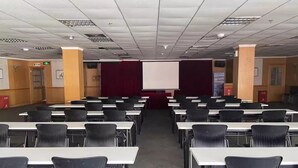 Une salle de conférence avec des rangées de tables et de chaises, un écran de projection et un rideau rouge en arrière-plan.