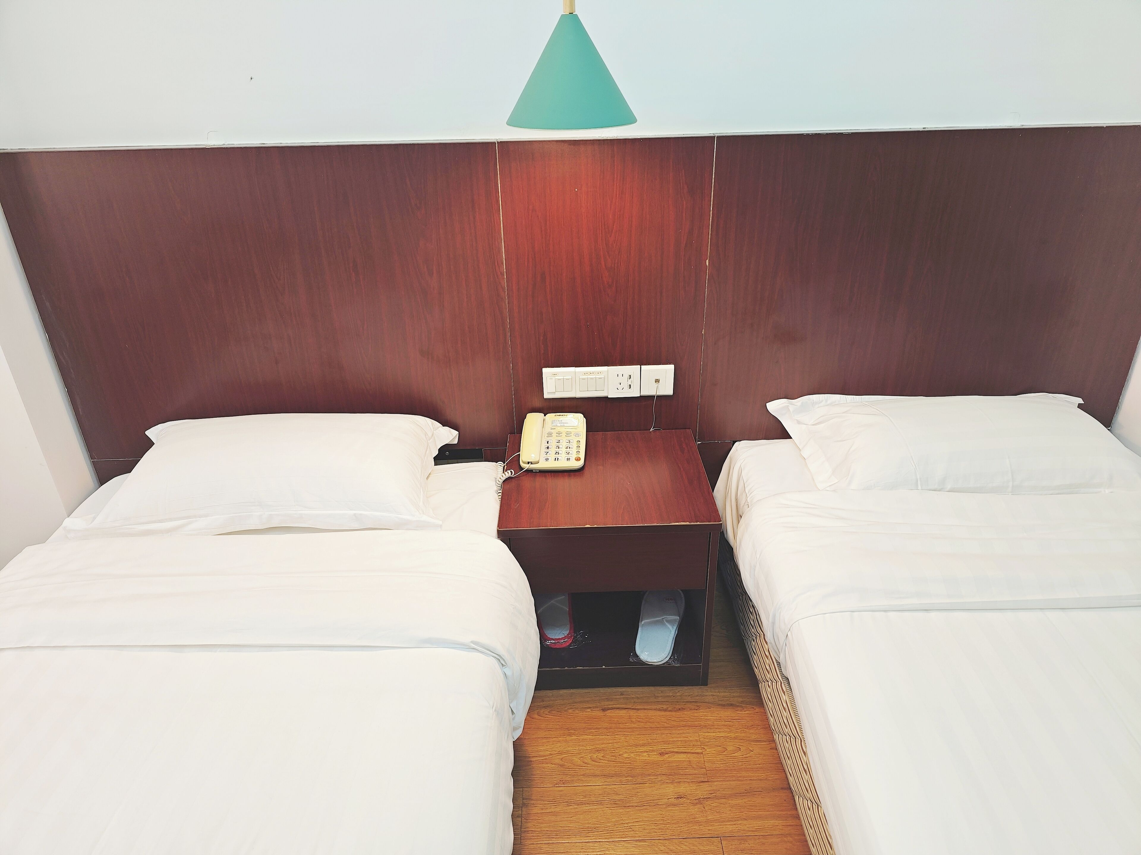 A hotel room with two single beds, a wooden headboard, a bedside table with a telephone, and a light fixture above.