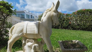A stone statue of a horse with a saddle, placed on a grassy area with a hedge and a building in the background.