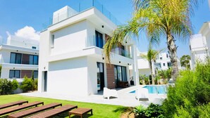 A modern two-story building with a swimming pool, outdoor seating, and lush greenery. - By the Beach Villa Natalya (Protaras)
