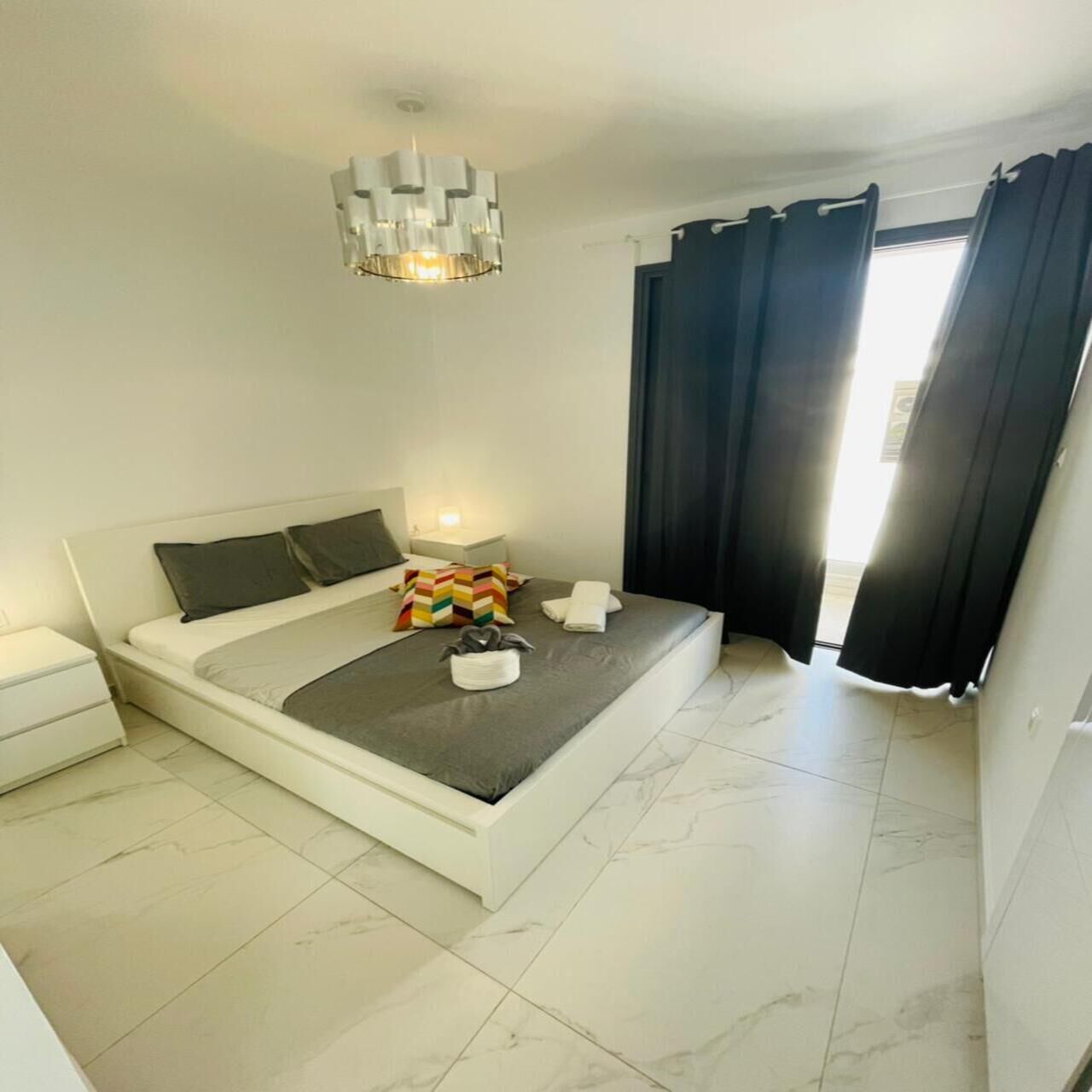 Une chambre moderne avec un grand lit, une table de chevet, un luminaire suspendu et une vue sur l’extérieur par une fenêtre agrémentée de rideaux.