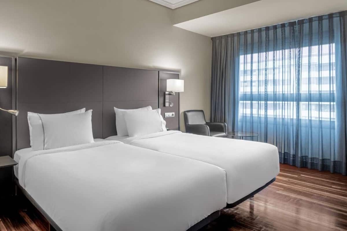 Une chambre d’hôtel avec deux lits, une table de chevet, une lampe, une chaise et une grande fenêtre avec des rideaux.