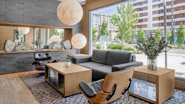 Un hall d’hôtel moderne avec un mur en briques grises, une grande fenêtre donnant sur la ville, un canapé gris, une table basse en bois, un fauteuil en cuir et une suspension ronde.