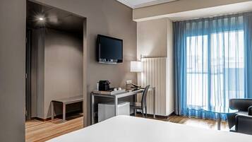Habitación de hotel con una cama, un escritorio con una silla, un televisor en la pared, una lámpara y un ventanal con cortinas.