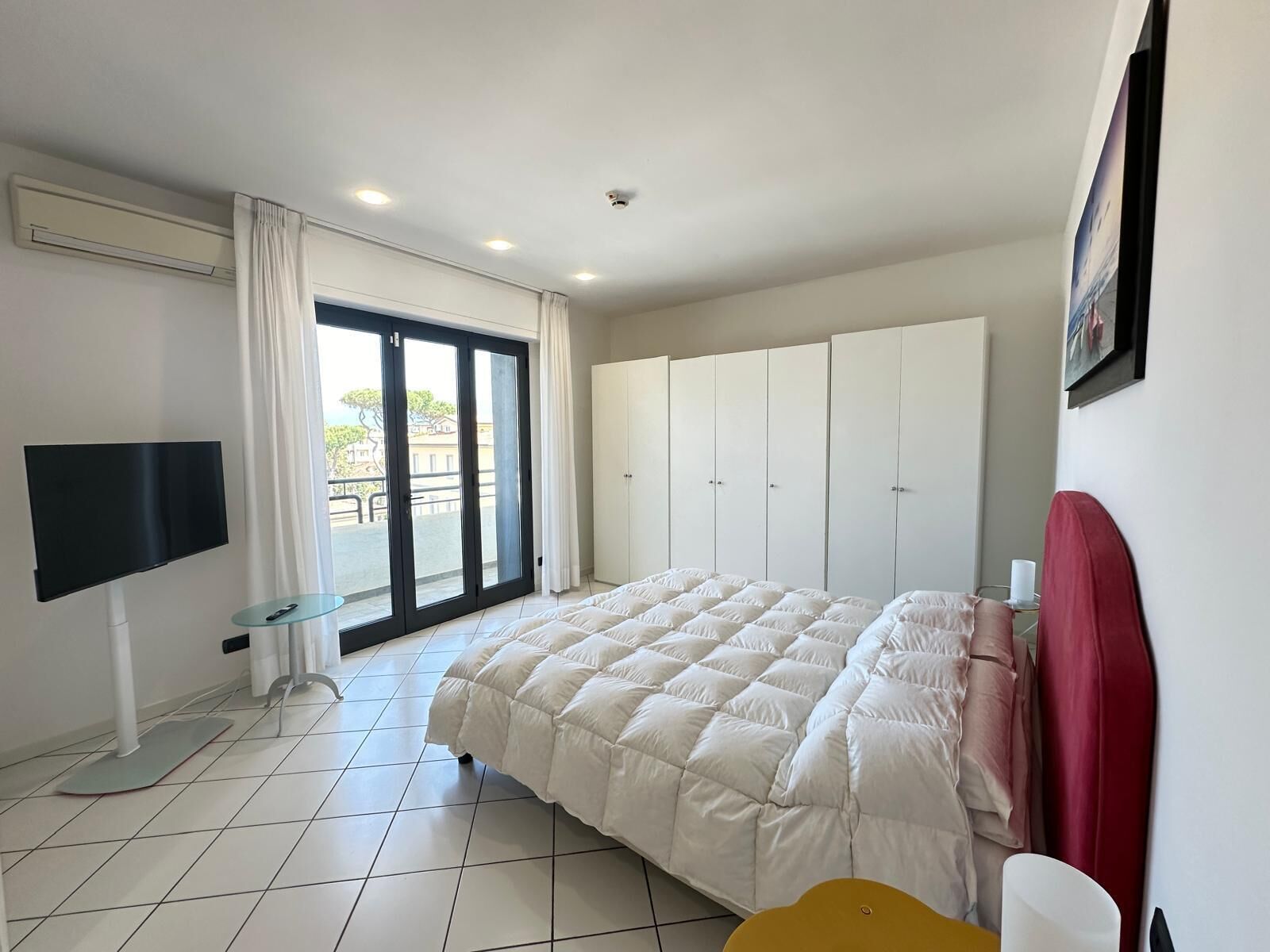 A modern bedroom with a large bed, a flat-screen TV, a sliding glass door to a balcony, and a wardrobe.