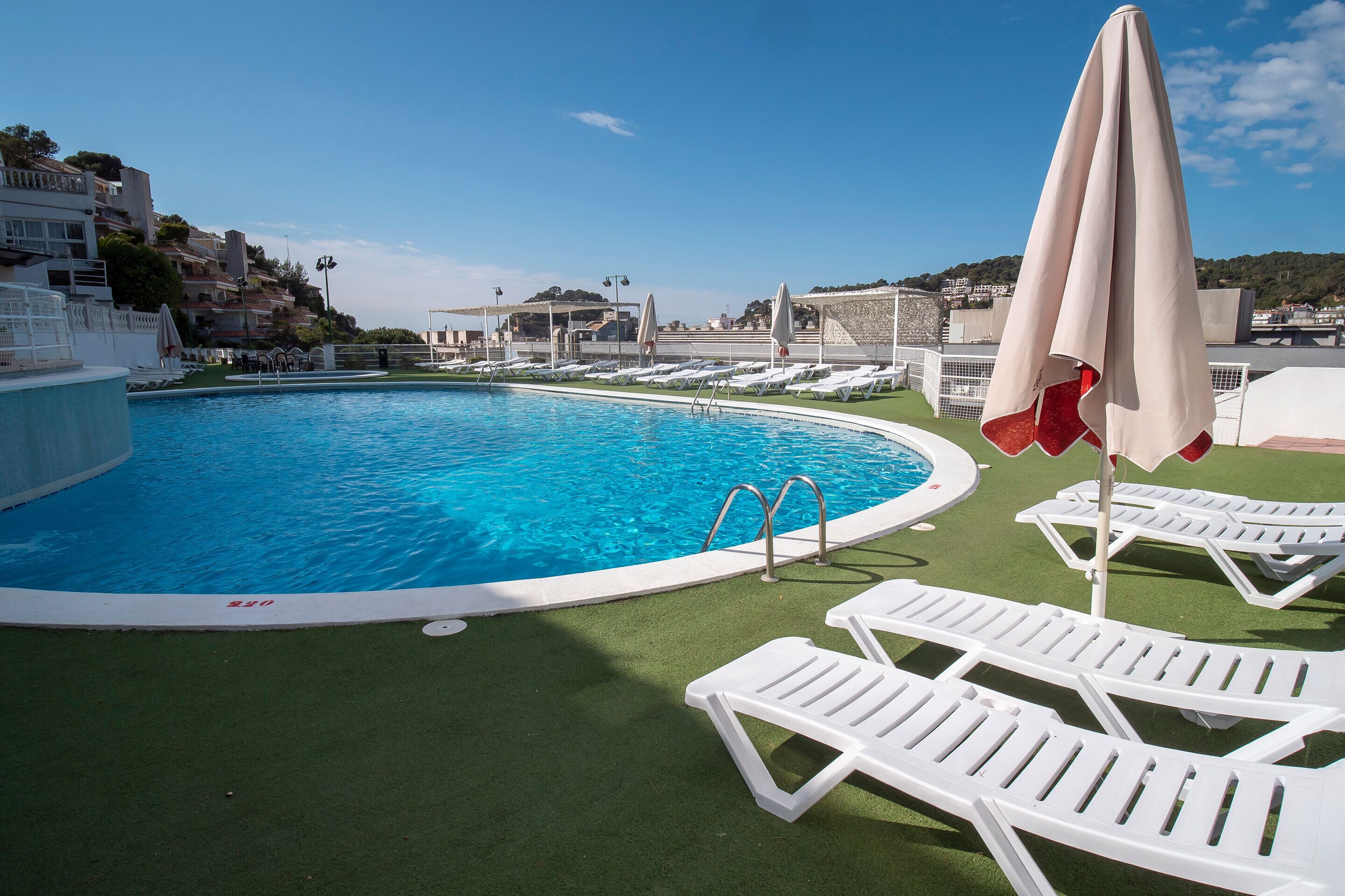 Une piscine bordée de chaises longues blanches et d’ombrelles, entourée d’une pelouse verdoyante et de bâtiments en arrière-plan.