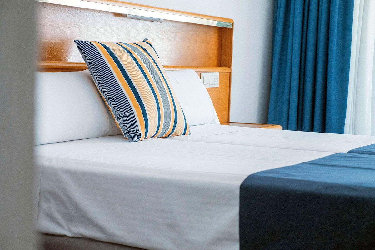 Una cama bien hecha con ropa blanca y una almohada a rayas, un cabecero de madera y una cortina azul.