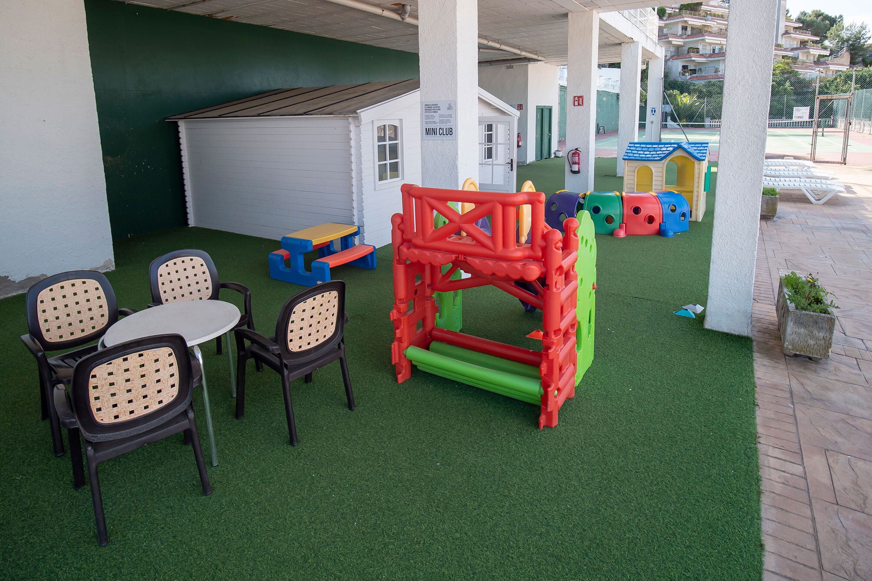 Un área al aire libre con mesas y sillas, un parque infantil y un cartel que indica 'mini club'.