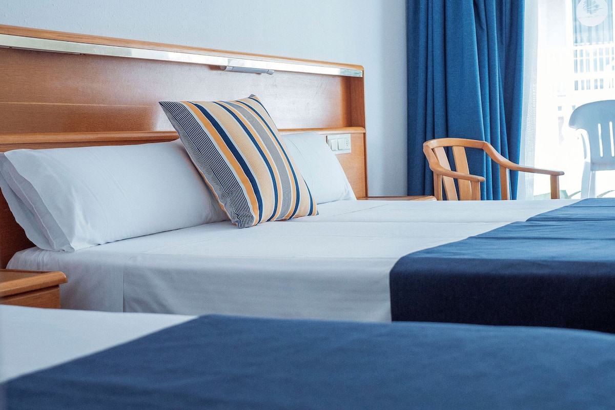 Habitación de hotel con cabecera de madera, cama con ropa blanca y almohadas a rayas, colcha azul, silla de madera y ventana con cortinas azules.