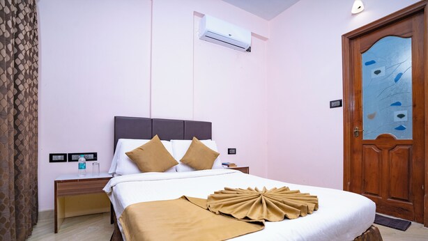 A neatly made bed with a headboard, a bedside table with a bottle and glasses, and a wooden door with a glass panel. - SS Mystic Apartotel (Bengaluru)