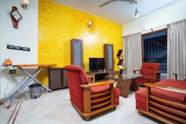 Una sala de estar con pared amarilla, televisor, sillas rojas y una mesa de centro de vidrio.