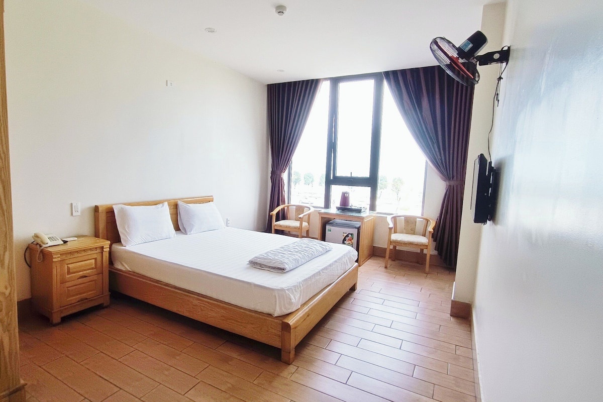 A hotel room with a wooden bed, a desk with a chair, a small table, and a fan.