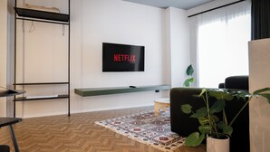 A modern living room with a flat-screen TV mounted on the wall, a wooden herringbone floor, a patterned rug, a black sofa, a small side table, and a potted plant. - Muz Studios (Tirana)