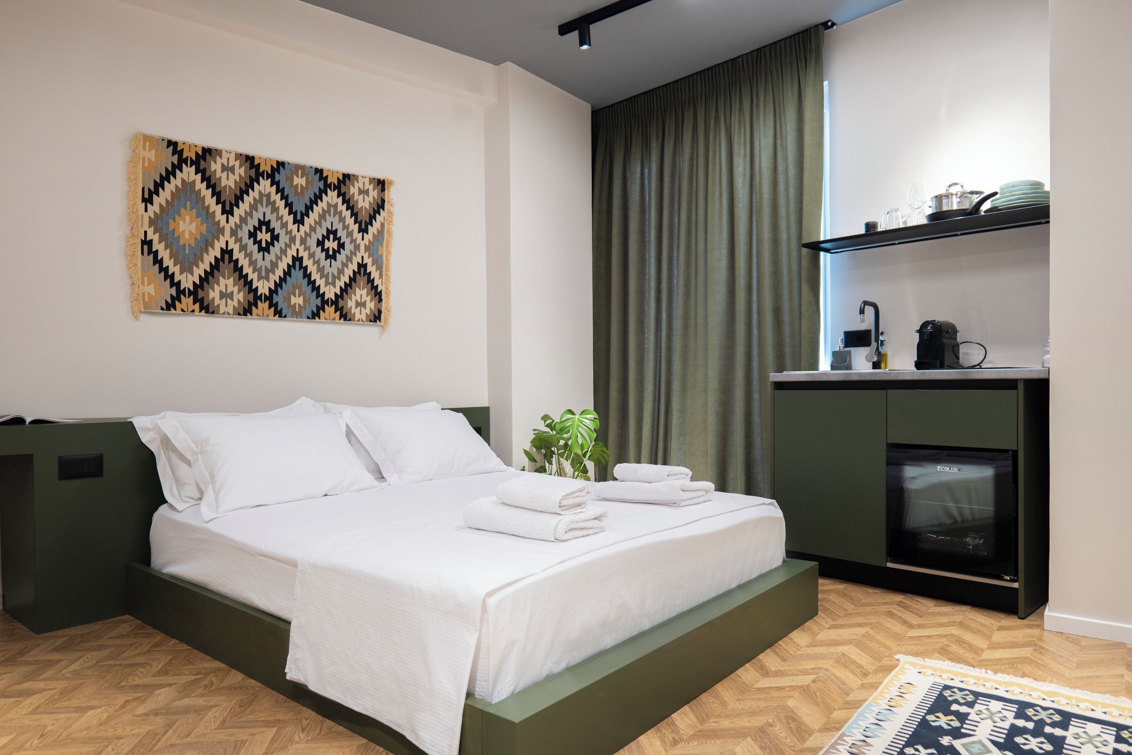 Quarto moderno com cama, mesa de cabeceira verde, obra de arte na parede, área de cozinha com fogão e janela com cortinas.