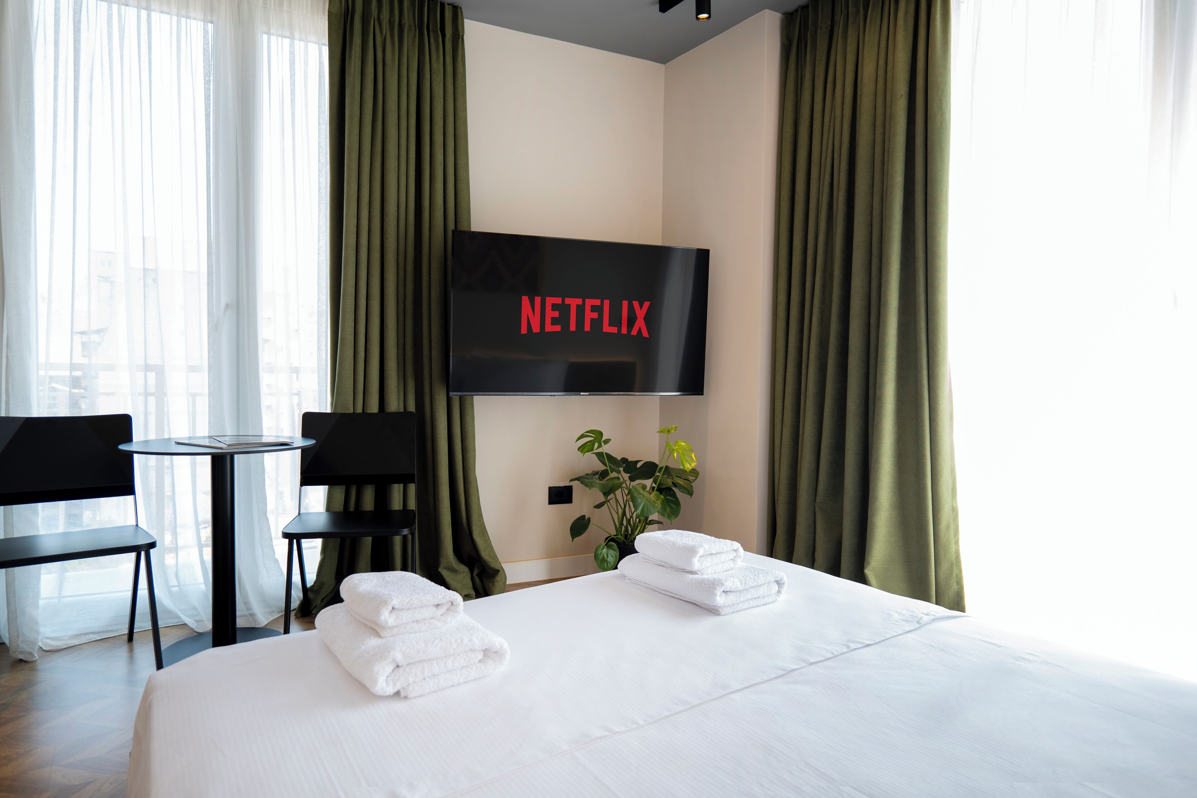A hotel room with a bed, a TV mounted on the wall, a small table, two chairs, and a potted plant.