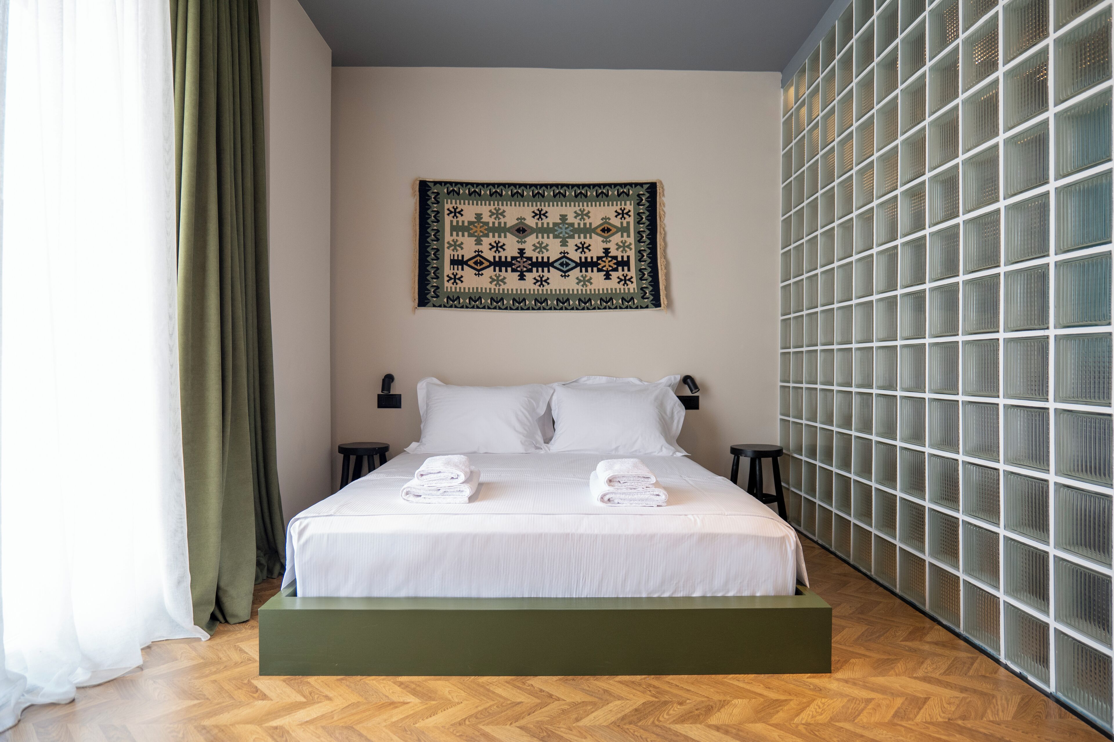 Una camera d'albergo moderna con un letto grande, una pedana verde, un arazzo appeso al muro e un'ampia finestra con tende.