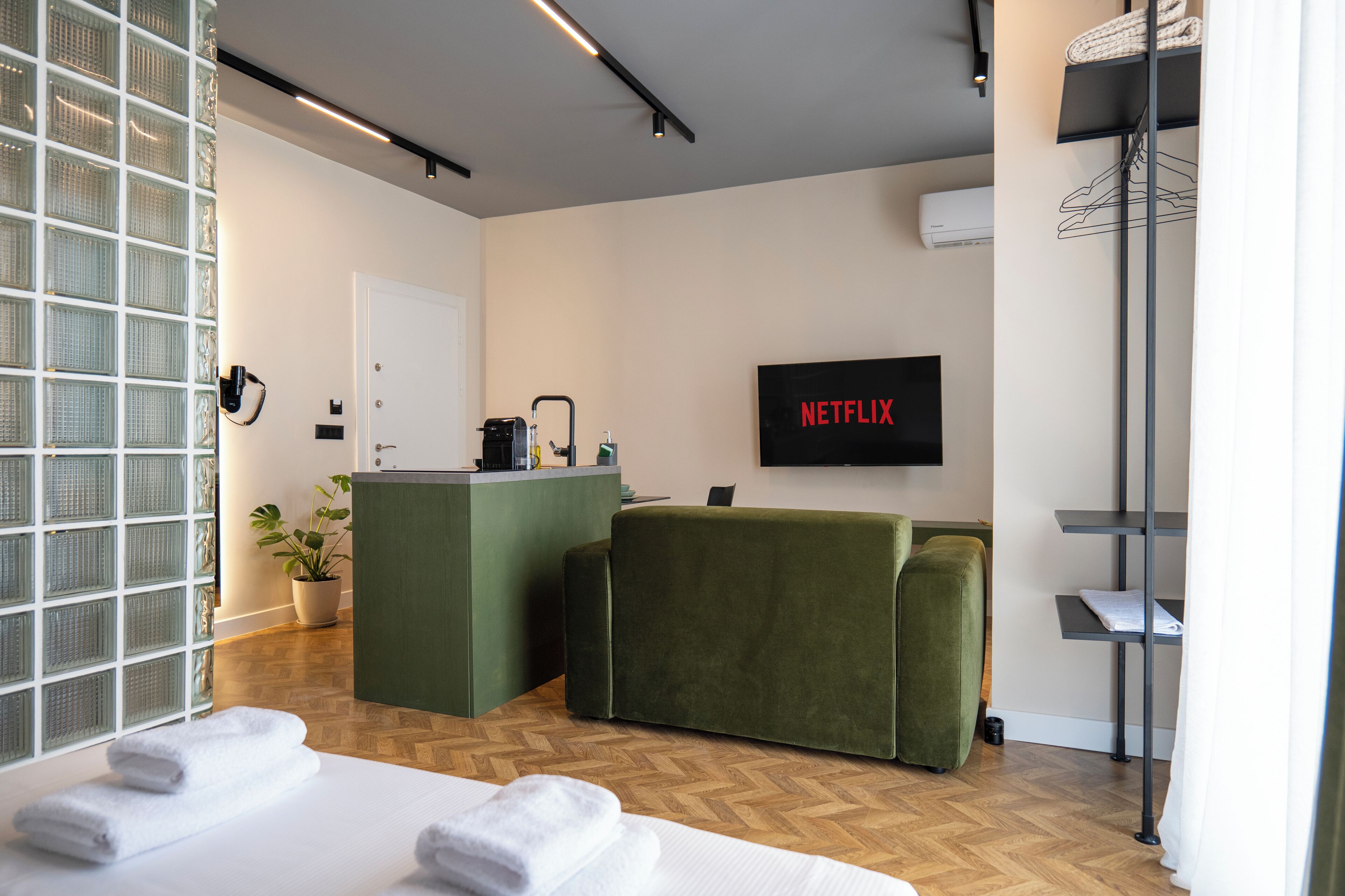 A modern hotel room with a reception desk, a green sofa, a bed with white linens, and a wall-mounted TV displaying 'NETFLIX'.