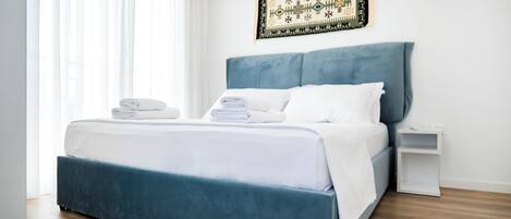 Un dormitorio moderno con una cama grande, toallas blancas dobladas con cuidado y un cabecero azul, sobre un piso de madera.
