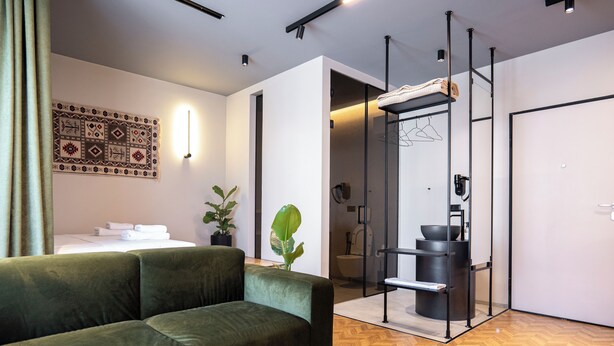 A modern hotel room with a green sofa, a patterned wall hanging, a plant, and a minimalist bathroom with a shower and a towel rack. - Muz Studios (Tirana)