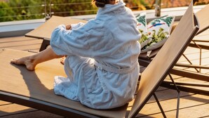 Eine Person sitzt auf einer Liege auf einem Balkon mit Blick auf die Küste.