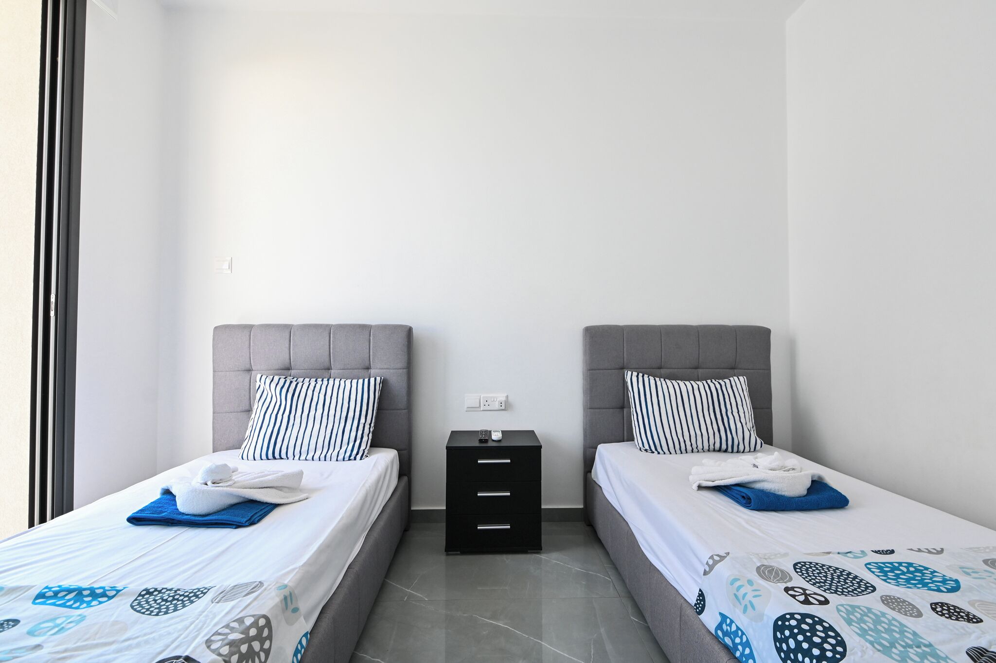 Une chambre double avec deux lits, chacun doté d’une tête de lit grise, d’une table de chevet noire avec des tiroirs et d’un mur blanc.