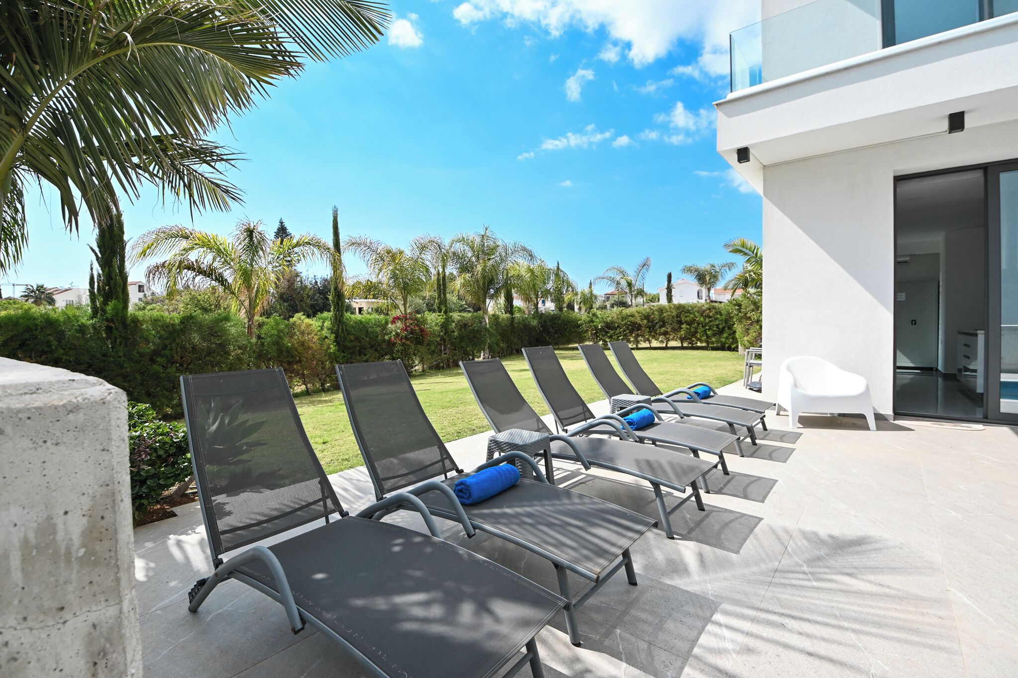A modern outdoor seating area with lounge chairs and a white chair, surrounded by greenery and a clear sky.