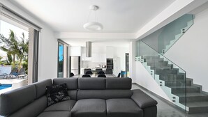 Ein modernes Wohnzimmer mit einem grauen Ecksofa, einer Glasstairnase und Blick auf die Küche und den Essbereich.