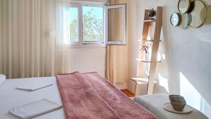 Quarto com cama, mesa de cabeceira, manta rosa, janela com cortinas e prateleira com objetos decorativos.