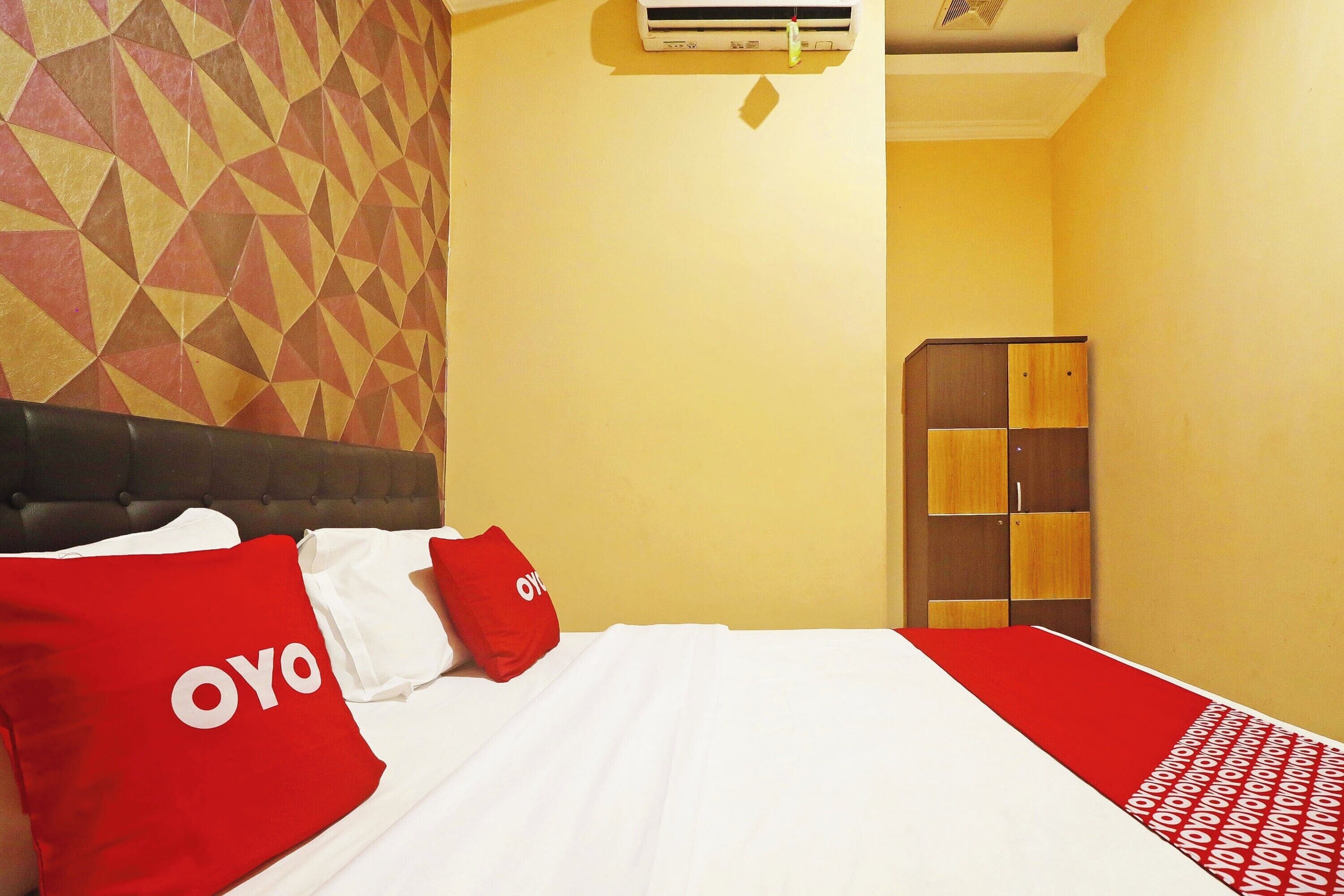 A hotel room with a patterned wall, a bed with red and white bedding, and a wooden wardrobe.