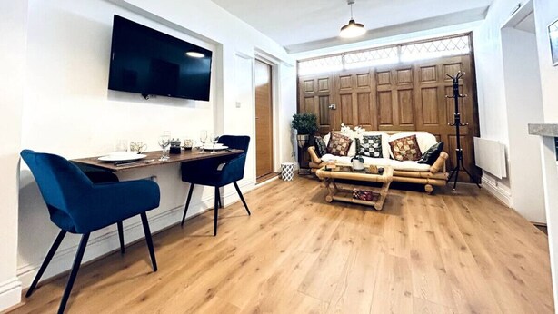Ein modernes Wohnzimmer mit Fernseher, einer Essecke mit zwei blauen Stühlen, einer Holzsofa mit gemusterten Kissen und Holzboden.