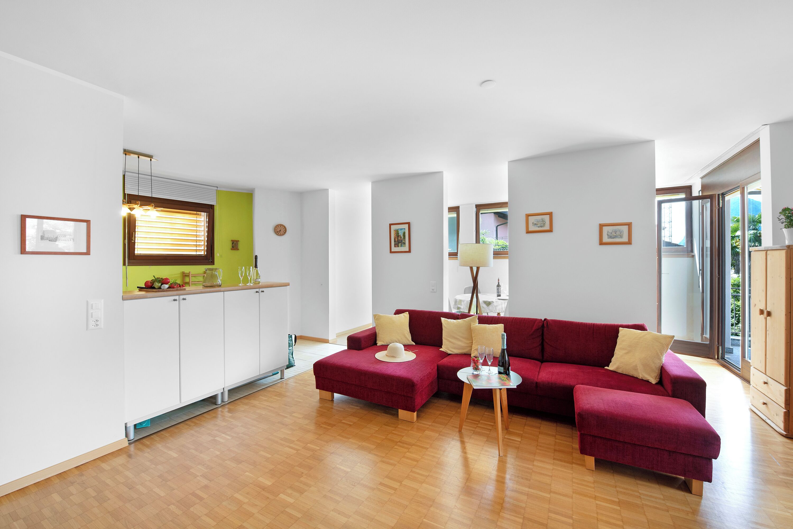 A modern living room with a red sectional sofa, a small coffee table, and a kitchen area with white cabinets and a green accent wall.