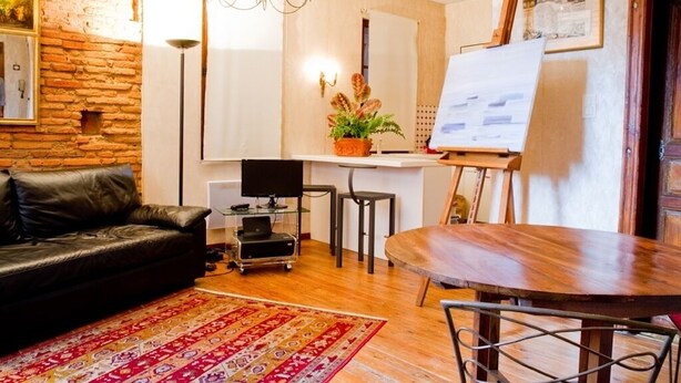 A living room with a black leather sofa, a wooden dining table, a red patterned rug, a television on a stand, a whiteboard, and a potted plant. - Bel Appartement T1bis de Caractère Jardin Royal. Quartier des Carmes (Toulouse)