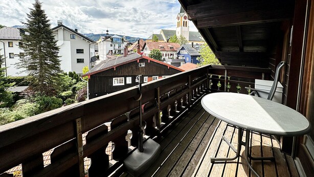 A wooden balcony with a table and chairs, overlooking a town with buildings and a church. - Calendula vacation apartment (Oberstaufen)