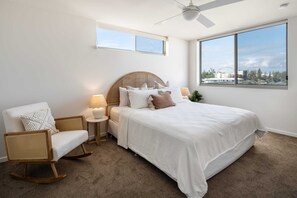 A bedroom with a bed, a rocking chair, a nightstand, and a window with a city view.