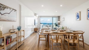 Un comedor moderno con una mesa y sillas de madera, una estantería y vistas al océano a través de amplias ventanas.