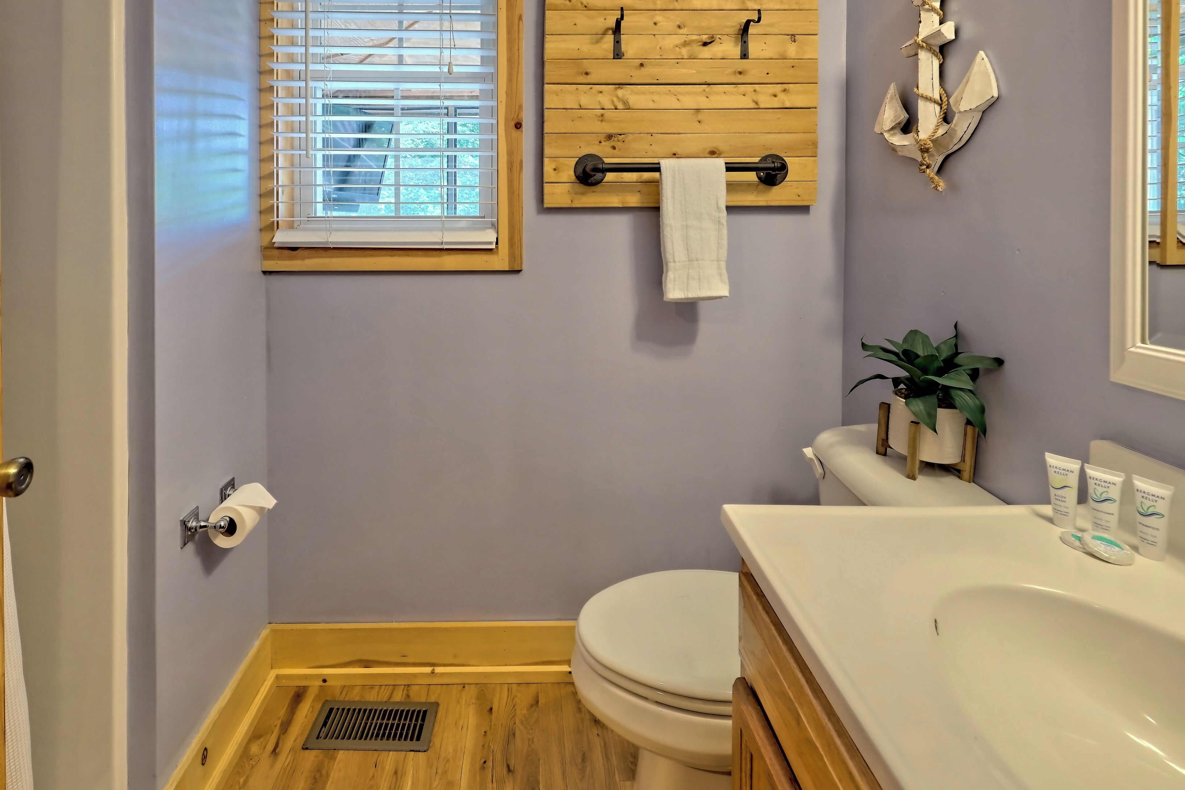 A bathroom with a wooden floor, a white toilet, a sink with toiletries, and a window with blinds.