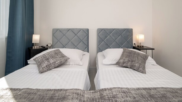 A hotel room with two beds, each with a grey headboard and white bedding. There are bedside tables with lamps and a small clock on each.