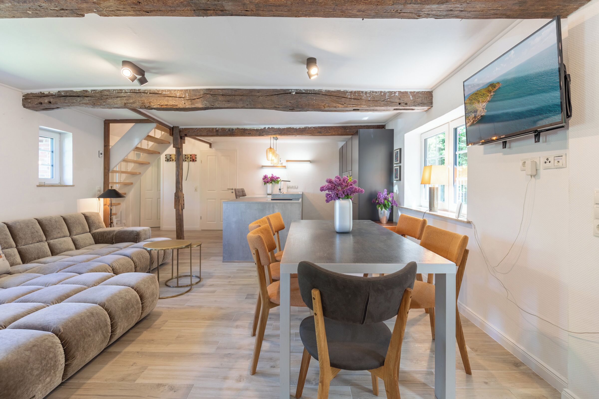 Ein modernes Wohnzimmer mit einer Couch, Esstisch und Stühlen. Im Hintergrund ist eine Küche erkennbar, mit einer Treppe, die eine weitere Ebene hinaufführt.