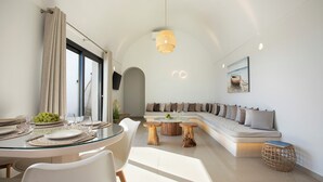 A modern living room with a glass dining table, white chairs, a wooden coffee table, and a built-in sofa with cushions. - Vinebranch Villas (Santorini)
