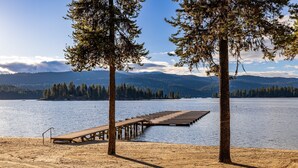 Vista su un lago, con un pontile di legno che si protende nell'acqua, circondato da alberi e montagne sullo sfondo.