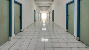 Un passadís llarg i estret amb portes blaves als costats, que condueix a habitacions individuals. El terra està emblenat i el passadís està ben il·luminat amb llums al sostre.