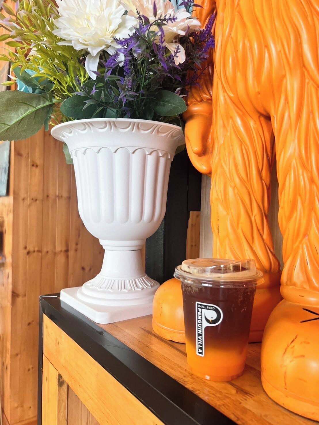 A white ceramic vase with flowers, a plastic cup with a label, and a pumpkin decoration on a wooden surface.
