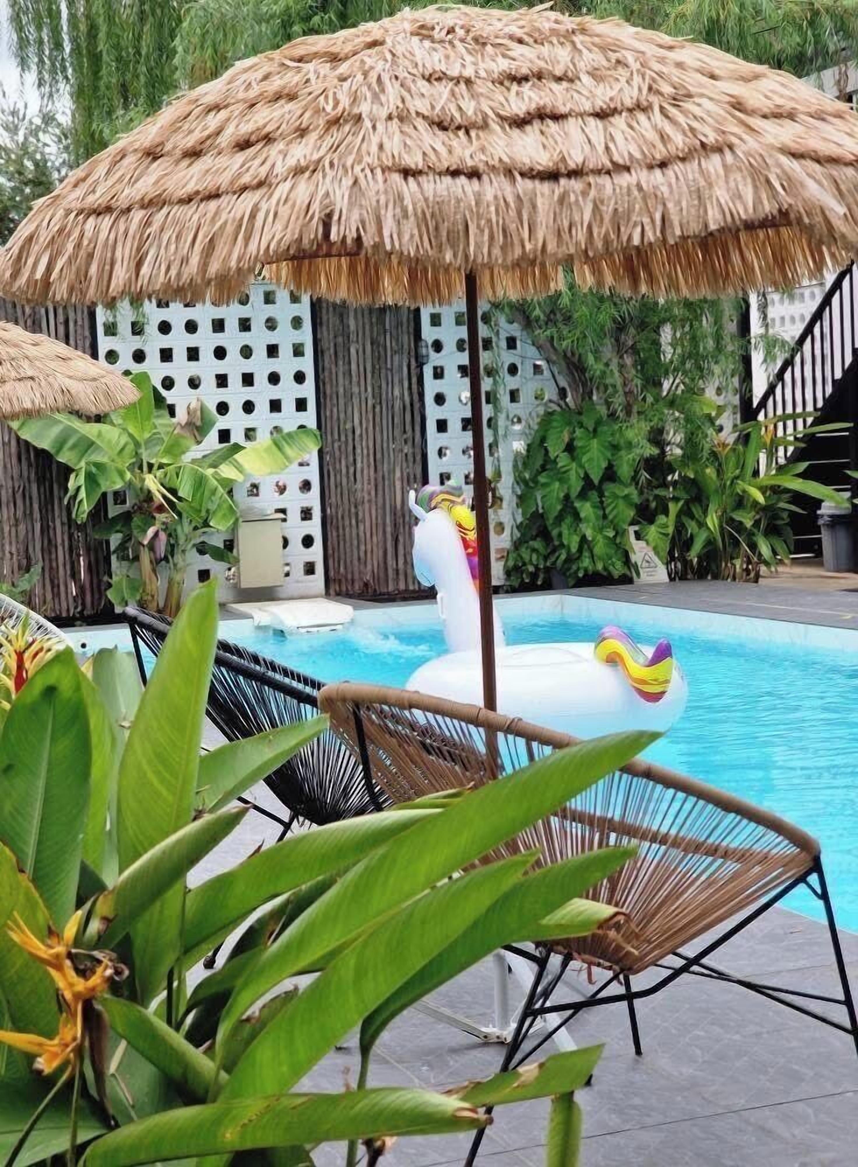 Área de piscina com guarda-sol de palha, espreguiçadeiras e plantas tropicais.