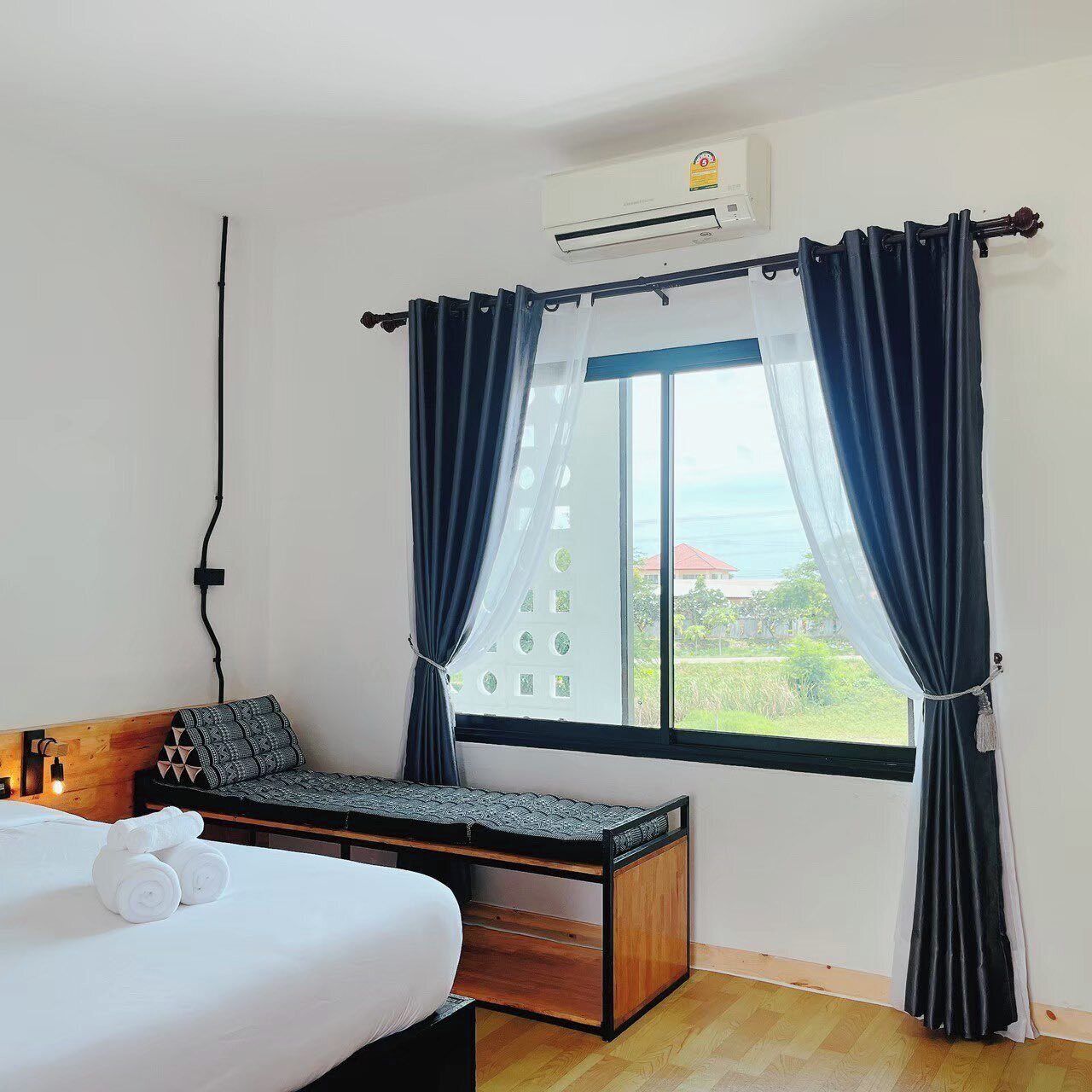 Un dormitorio con cama, un banco de madera, una ventana con cortinas, un equipo de aire acondicionado y vistas al exterior.