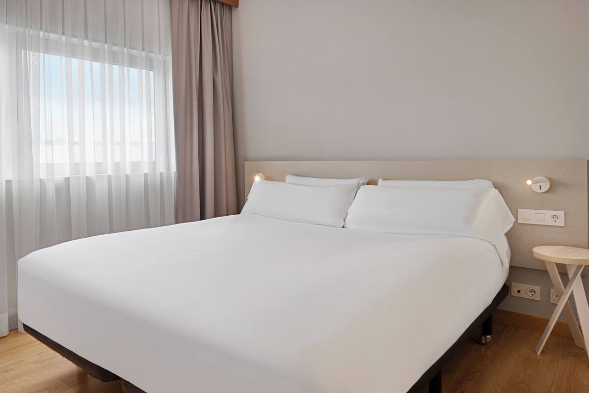 A neatly made bed with white linens in a room with a wooden floor, a small bedside table, and a wall-mounted light.