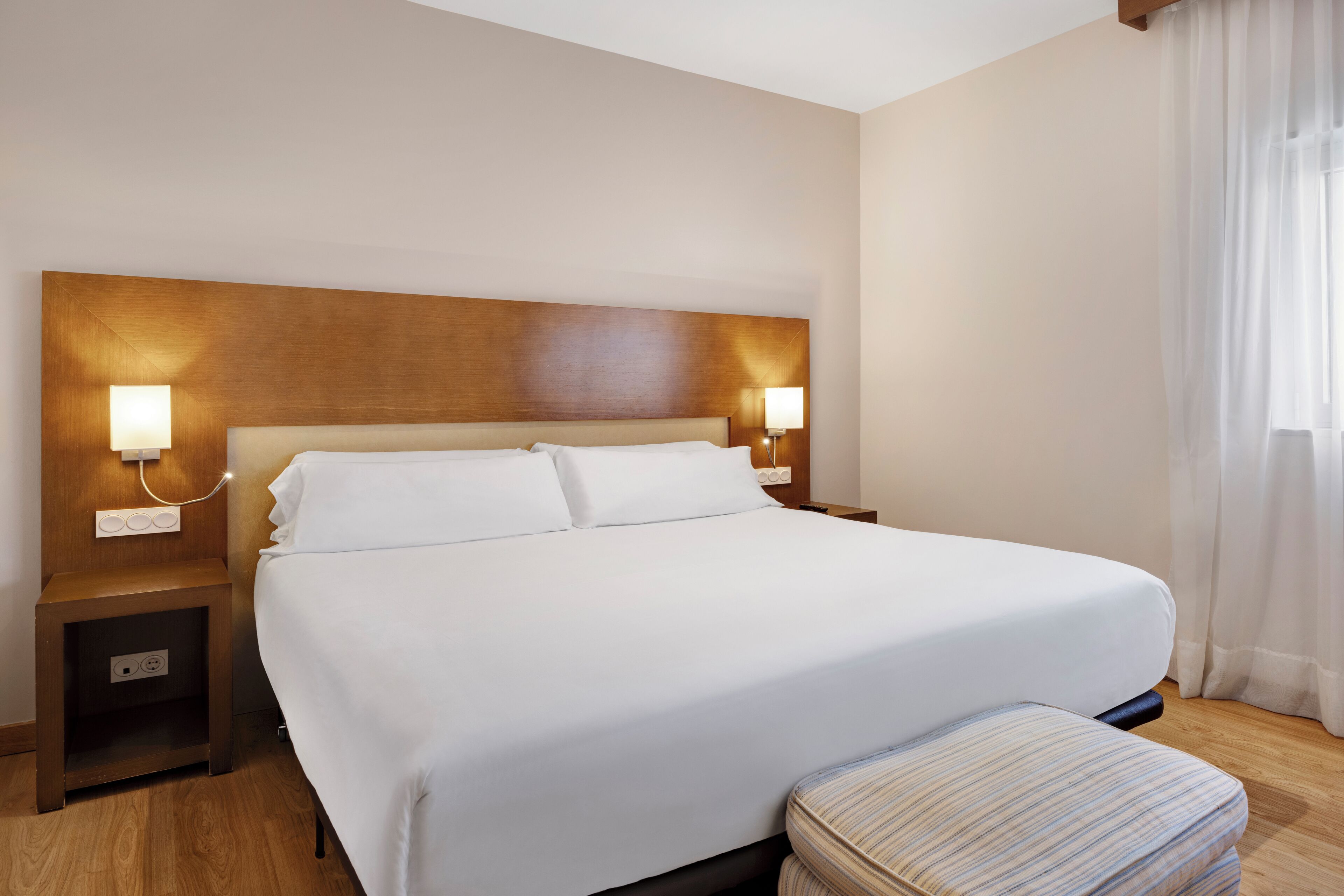 A hotel room with a large bed, two bedside tables with lamps, a wooden headboard, and a window with curtains.