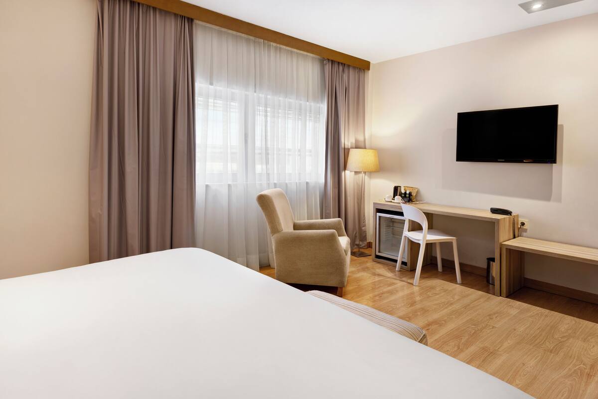 A hotel room with a large bed, a desk with a chair, a TV mounted on the wall, and a window with curtains.