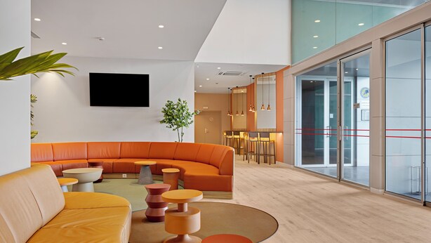 A modern lobby with a curved orange sofa, round wooden tables, and a large flat-screen TV mounted on the wall. - B&B Hotel Logroño Las Cañas (Viana)