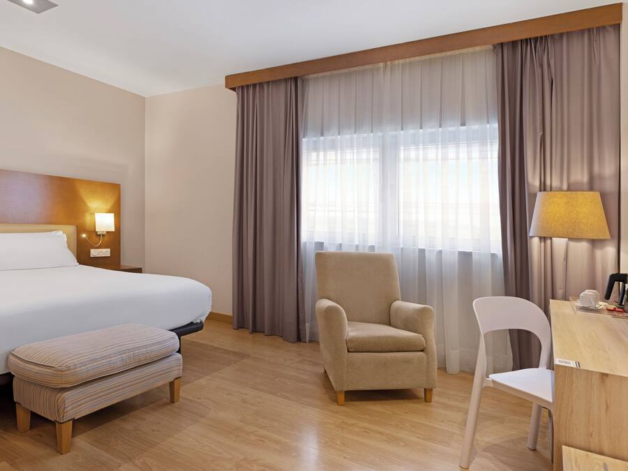 Habitación de hotel con una cama grande, un escritorio de madera, una silla, un sillón pequeño y una ventana con cortinas.
