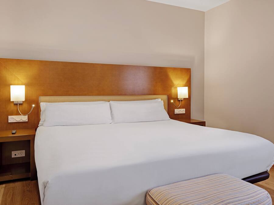 Una habitación de hotel con una cama grande, lámparas de noche, una mesita de noche y un cabecero de madera.